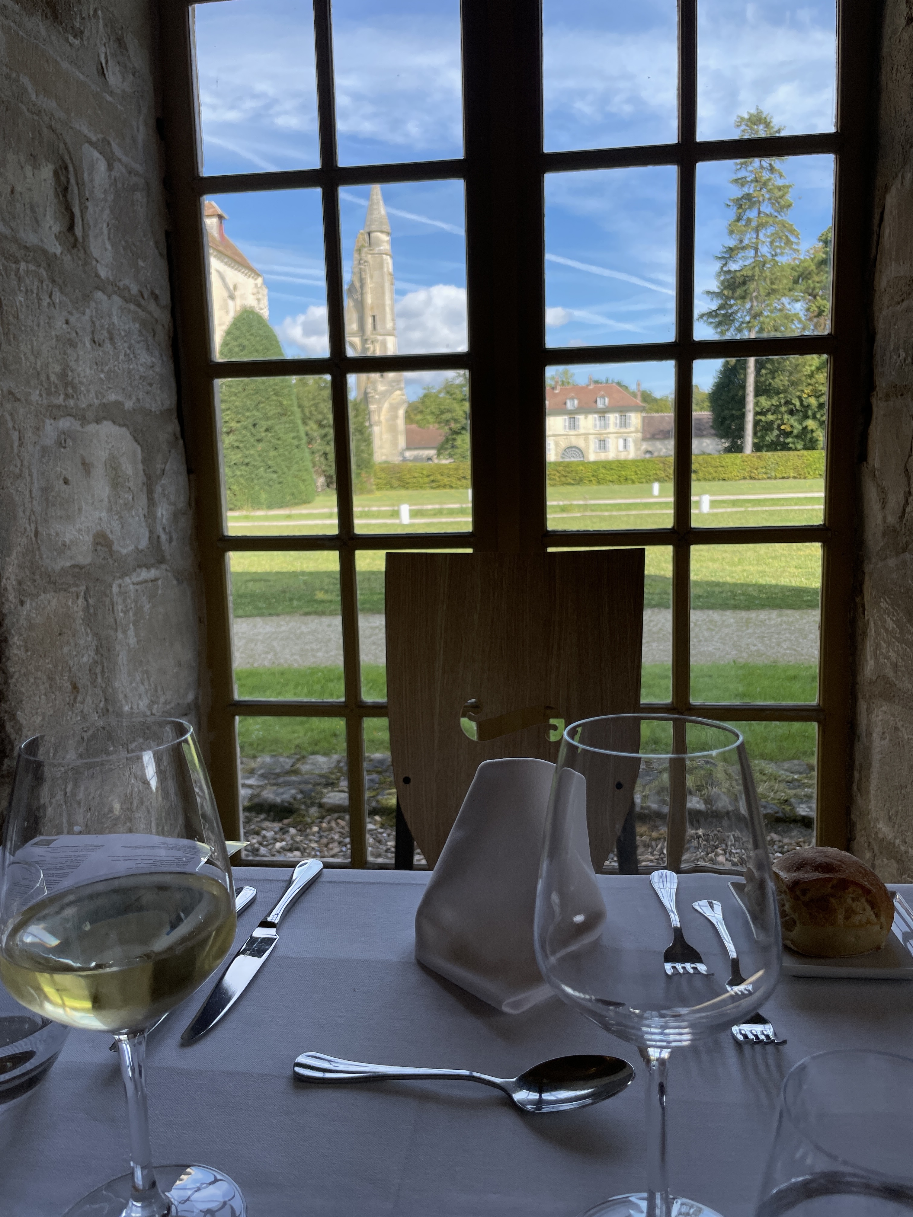 Vue depuis une table de restaurant avec des verres et couverts, montrant une fenêtre avec des bâtiments historiques et un paysage verdoyant à l'extérieur.
