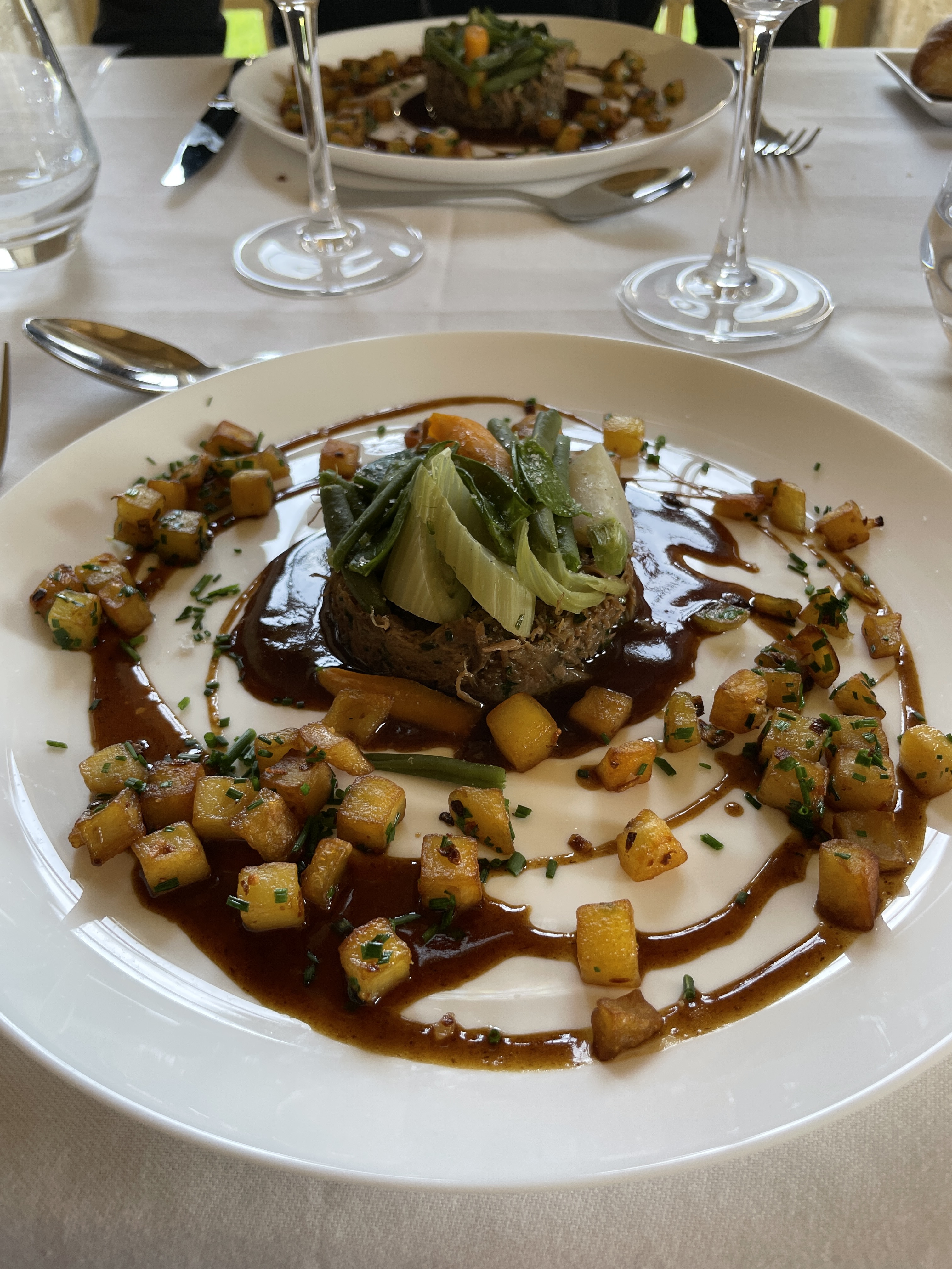 Un plat gastronomique avec une portion de viande accompagnée de légumes, de pommes de terre sautées et d'une sauce, servi dans une assiette blanche.