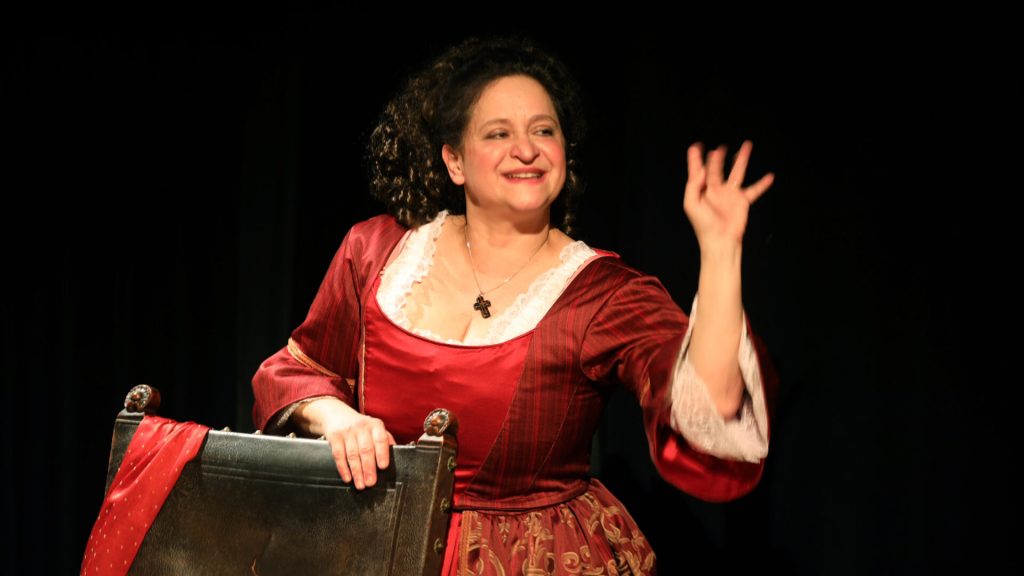 Une femme souriante en costume d'époque lève la main pour saluer, devant un fond noir.