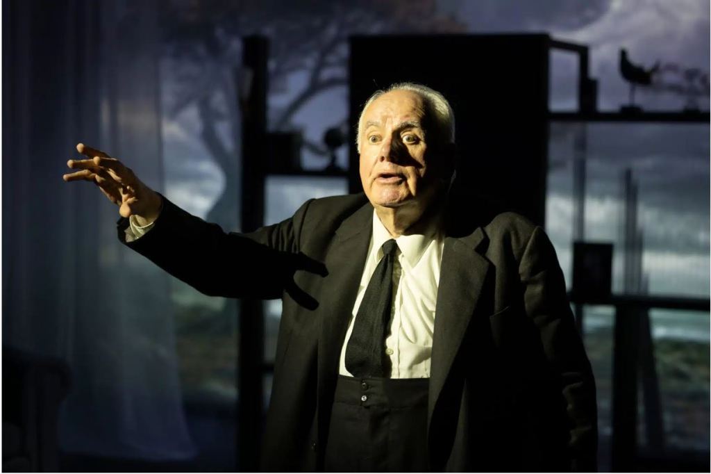 Un homme âgé, habillé en costume noir, s'exprimant avec passion dans un environnement dramatique.