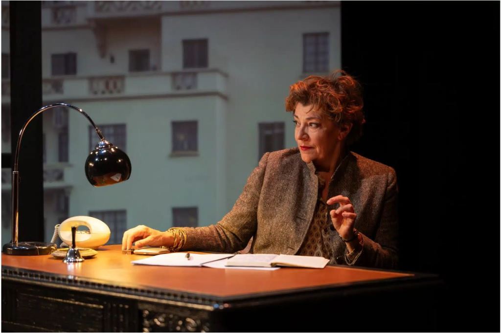 Une femme assise à un bureau avec une lampe de bureau, un téléphone et des notes, devant un fond représentant un bâtiment.