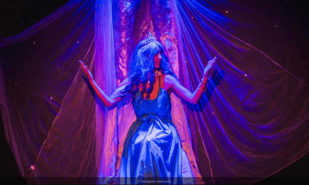 Une artiste en costume de scène avec une robe bleu clair et des cheveux longs, posant derrière un rideau translucide illuminé par des lumières violettes.