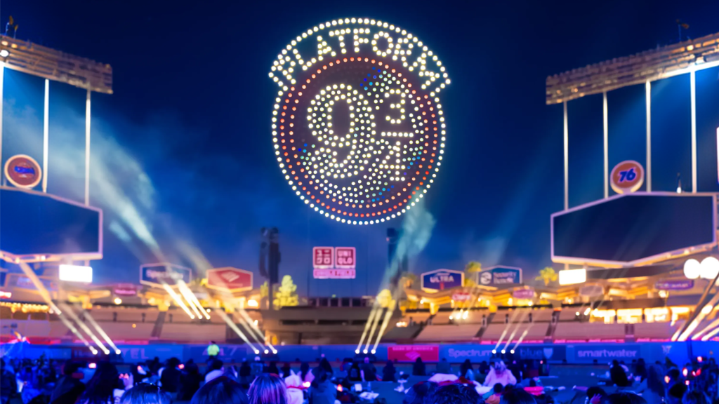 A night view of a stadium with a large illuminated sign reading 'Platform 9 3/4' in the sky, surrounded by colorful lights and a crowd of people in the foreground.
