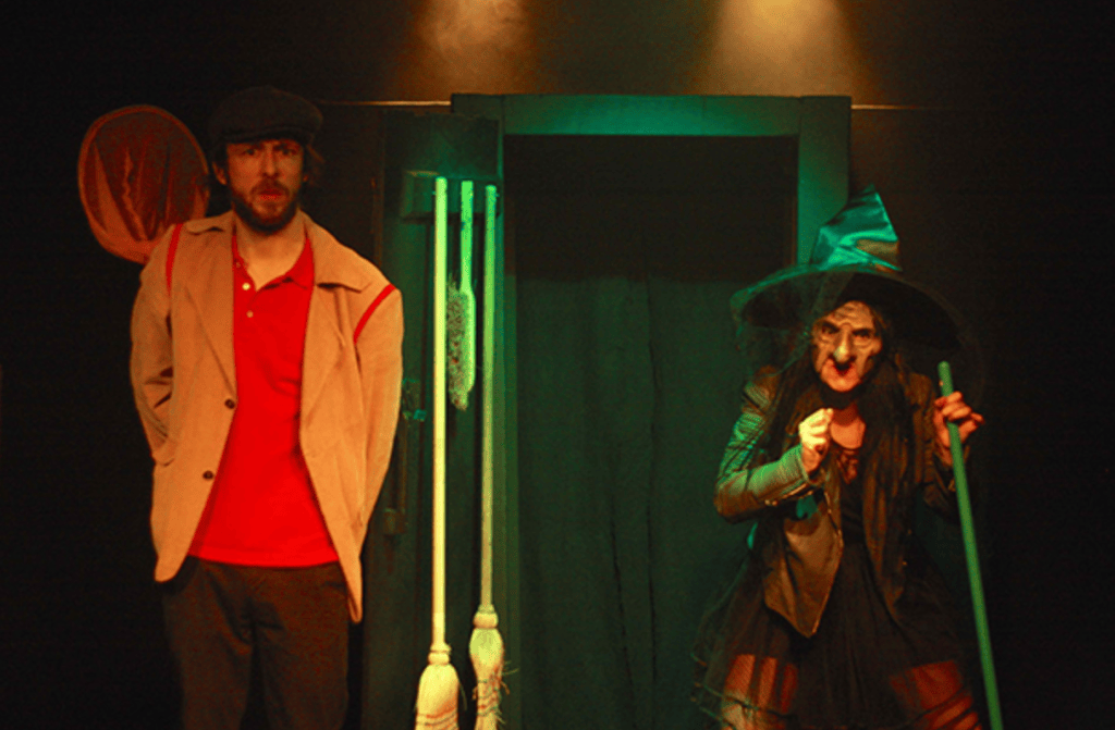 Une scène de théâtre avec un homme au visage sérieux portant une veste claire et un polo rouge, et une femme habillée en sorcière brandissant un bâton, dans une ambiance sombre et mystérieuse.