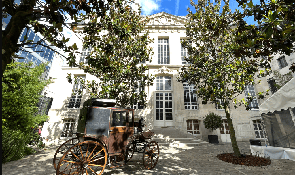 Un bâtiment historique avec des arbres, une cour pavée et une calèche ancienne à l'avant.