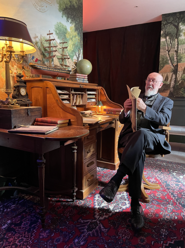 Un homme âgé assis à un bureau en bois, lisant un document. La pièce est décorée avec un lampadaire, un globe et des livres, avec une tapisserie d'arrière-plan représentant des voiliers.