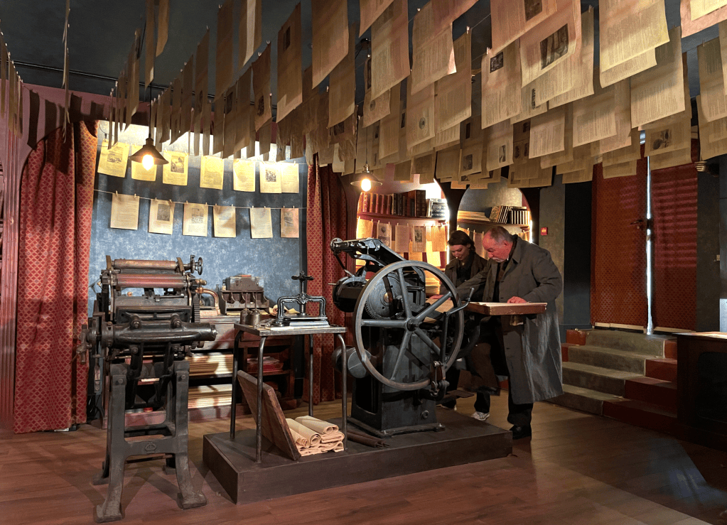 Un atelier d'imprimerie avec des machines anciennes et des papiers suspendus au plafond. Deux hommes examinent des documents sur une table.