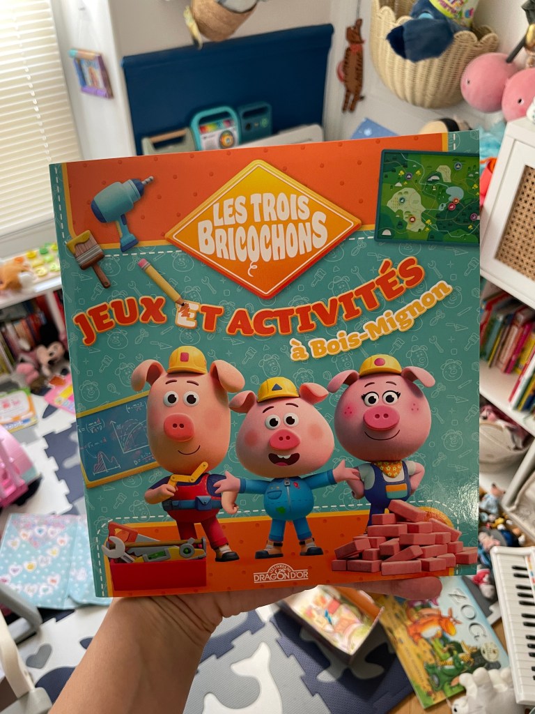 Couverture d'un livre intitulé « Les Trois Bricochons : Jeux et Activités à Bois-Mignon », présentant trois cochons animés au style cartoon, tenant des outils et entourés d'éléments colorés liés à l'activité.