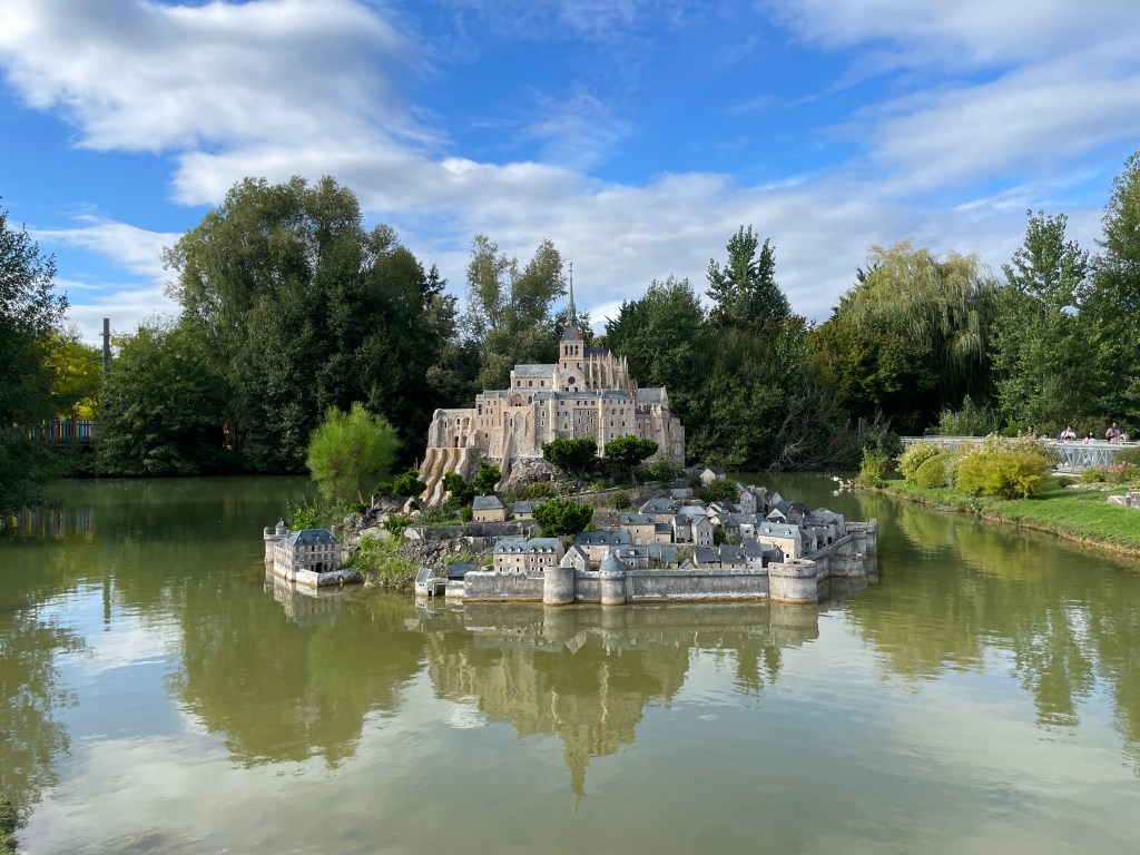 Maquette d'un château sur une île entourée d'eau, avec des arbres en arrière-plan et un ciel partiellement nuageux.