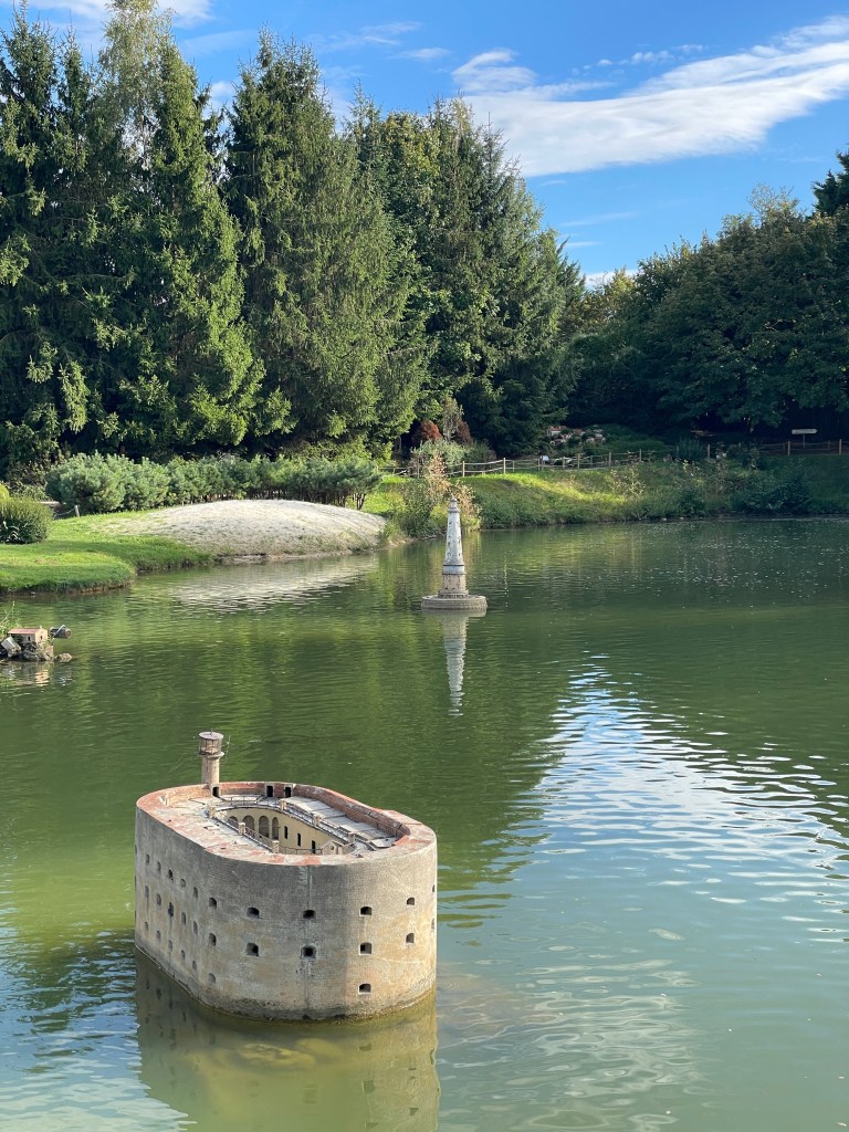 Un petit îlot cylindrique au milieu d'un lac, avec un édifice en pierre au fond et des arbres verdoyants tout autour sous un ciel bleu.