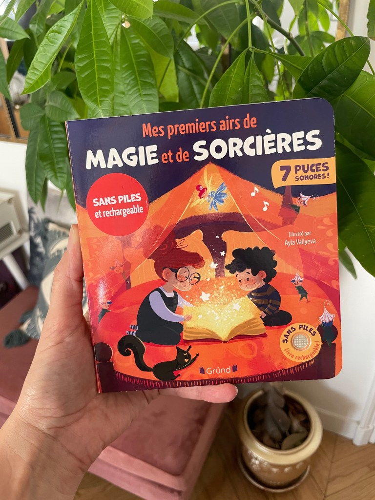 Un enfant tenant un livre intitulé 'Mes premiers airs de magie et de sorcières' avec des illustrations colorées, entouré de plantes d'intérieur.