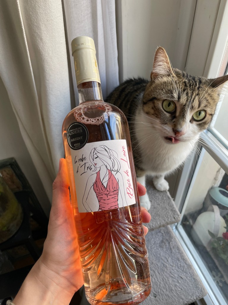 Une main tenant une bouteille de vin rosé avec une étiquette illustrée d'une femme, à côté d'un chat sur une fenêtre.